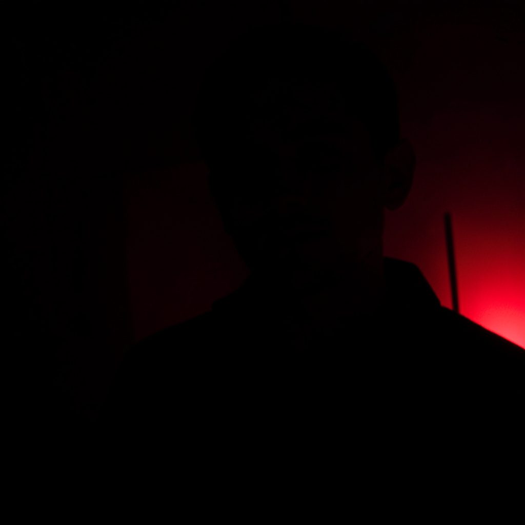 Dark, low-key photograph of a worn notebook page in deep shadow, with faint ink scribbles and a single warm reddish glow near the corner, conveying a quiet late-night mood.
